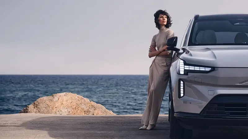 Žena stojaca pri Volvo EX90 pri morskom pobreží – prémiové elektrické SUV s pokojným škandinávskym charakterom